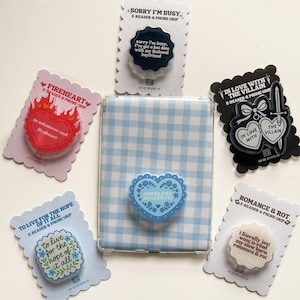 May include: An assortment of e-reader and phone grips with various designs and quotes. The grips feature heart, floral, and quote designs in colors like red, blue, and black. A blue and white gingham e-reader case is also present.