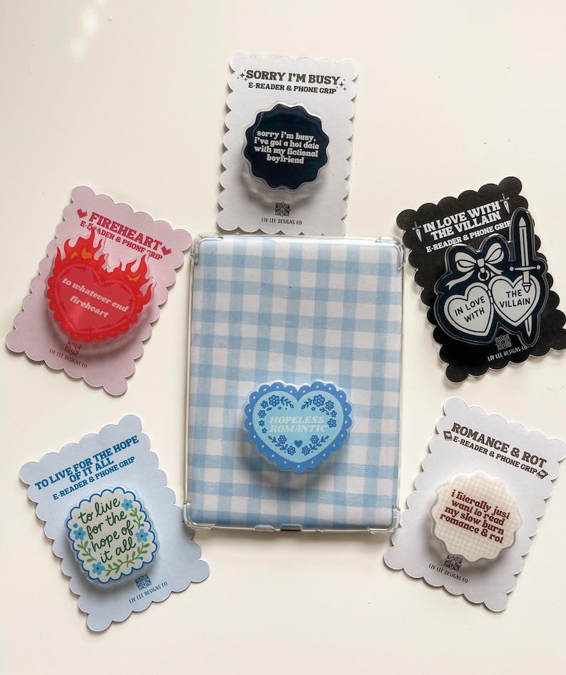 May include: An assortment of e-reader and phone grips with various designs and quotes. The grips feature heart, floral, and quote designs in colors like red, blue, and black. A blue and white gingham e-reader case is also present.