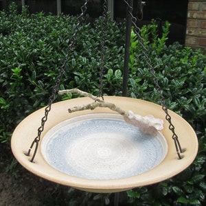 May include: A hanging bird bath with a light brown rim and a blue and white interior. The bird bath is suspended by black chains and features a decorative branch and a sculpted leaf-like element.