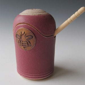 May include: A red ceramic honey pot with a bee design and a wooden honey dipper.