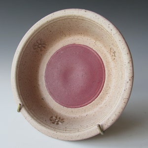 May include: A handmade ceramic bowl with a speckled beige exterior and a deep red center. The bowl features embossed floral designs around the rim and is supported by a brass stand. This decorative bowl is ideal for serving or display.
