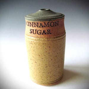 May include: A cylindrical ceramic shaker with a speckled tan body and a smooth, darker brown upper band featuring the impressed text CINNAMON SUGAR. The shaker has a dark green, slightly domed lid with a small circular opening at the very top center.