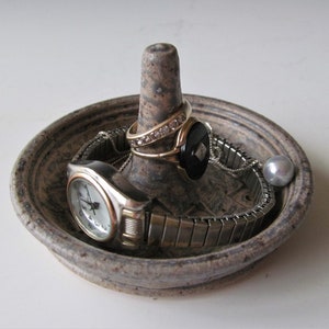May include: A ceramic ring holder with a raised center and a textured surface. The holder is filled with jewelry, including a watch, a ring with a black stone, and a ring with a row of diamonds.
