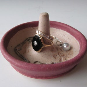 May include: A pink ceramic ring dish with a white center and a small white cone in the center. There are three pieces of jewelry on the dish: a black ring with a diamond, a gold ring with diamonds, and a silver chain with a pearl.