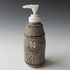May include: A ceramic soap dispenser with a white pump and a decorative design featuring a stylized flower and fan. The dispenser is brown and black with a textured surface.