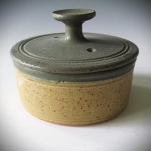 May include: A handmade ceramic butter dish with a lid. The dish has a speckled, light yellow base and a gray-green lid with a small handle. The lid has two small holes. The dish is round and has a rustic, handcrafted appearance.