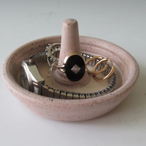 May include: A pink ceramic ring dish with a raised center cone. The dish is filled with jewelry, including a watch, a ring, and a pair of earrings.