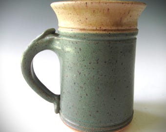 May include: Handmade ceramic mug with a speckled teal body and a tan rim. The mug features a large, curved handle and a slightly flared top. The base is a warm, earthy tone. This stoneware mug is perfect for coffee, tea, or any beverage.