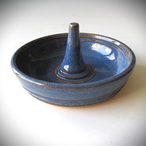 May include: A blue ceramic ring holder with a raised center cone. The holder has a glazed finish and a slightly textured surface.