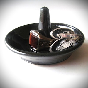 May include: A black ceramic ring holder with a central post and a shallow dish. A brown and silver ring sits on the post, and two silver car cufflinks rest in the dish. The ring holder is on a white surface.