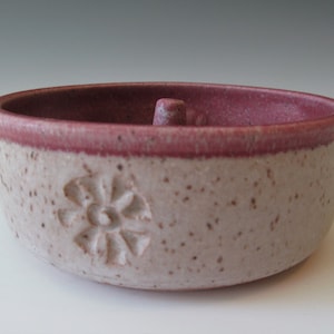 May include: A small, white ceramic bowl with a pink rim and a raised flower design. The bowl has a small, round knob in the center.