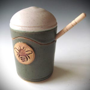 May include: A handmade ceramic honey pot with a lid and a wooden honey dipper. The pot is a muted green with a light beige lid and a carved bee emblem. The honey dipper is made of light-colored wood.