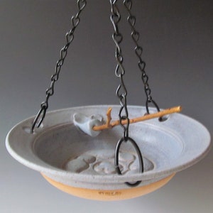 May include: A hanging ceramic bird feeder with a light blue bowl and a yellow-orange base. The bowl has a branch and bird design and is suspended by dark metal chains.