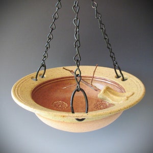 May include: A ceramic bird feeder with a brown and yellow glaze. The feeder has a wide rim and a small lip on one side. It is hanging from three black chains.