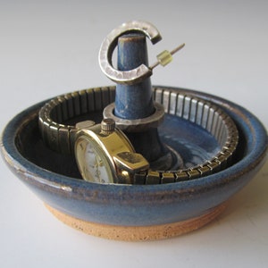 May include: A handmade blue ceramic jewelry holder with a watch, a silver hoop earring, and a silver ring. The watch has a gold face and a silver band. The jewelry holder has a ring holder and a small dish.