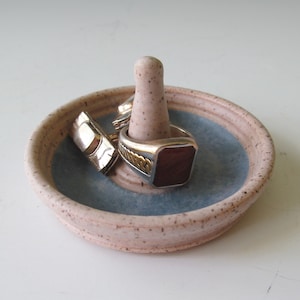 May include: A ceramic ring holder with a light blue glaze and a speckled beige finish. The holder has a raised center post for rings. Three rings are on the holder, one with a red stone.