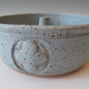 May include: A light blue ceramic bowl with a brown speckled glaze. The bowl has a raised design of a four-leaf clover on the side.