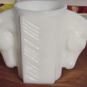 May include: A white, art deco-style vase with a cylindrical opening. The vase features two horse head sculptures on either side. The central panel has diagonal etched lines. The vase is made of opaque white glass.
