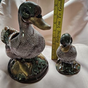 May include: Two ceramic duck figurines. The larger duck has a speckled body, a dark green head, and a brown beak. The smaller duck has a similar design. Both are on a base with green and brown accents. A yellow ruler is in the background.