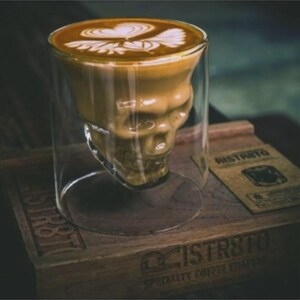 May include: A clear glass coffee mug shaped like a skull, filled with a latte featuring a heart-shaped design in the foam. The mug sits on a wooden tray with the word "Ristrto" printed on a small card.