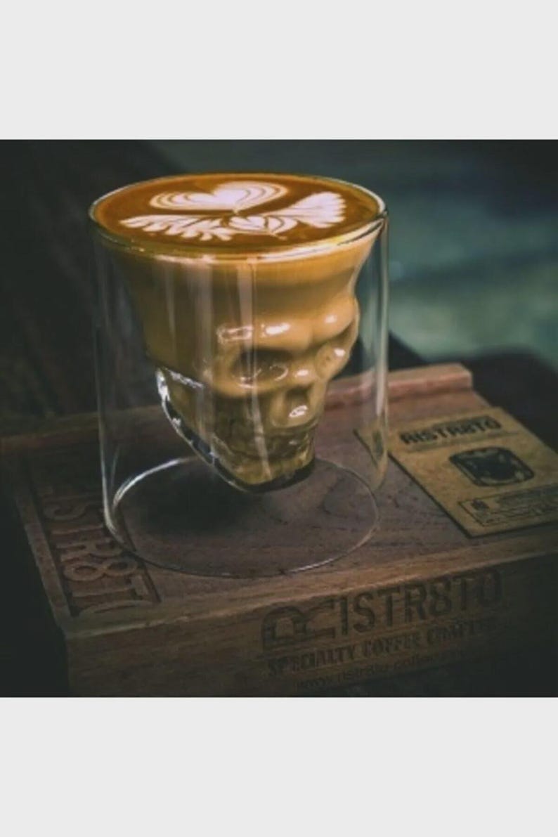 May include: A clear glass coffee mug shaped like a skull, filled with a latte featuring a heart-shaped design in the foam. The mug sits on a wooden tray with the word "Ristrto" printed on a small card.