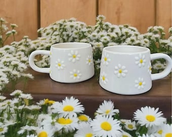 May include: Two white ceramic mugs with a daisy pattern. The mugs are sitting on a wooden surface in front of a bed of white daisies.