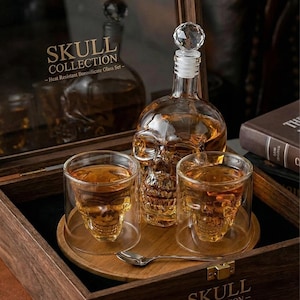Puede incluir: Un juego de vasos con forma de calavera que incluye una jarra con tapón de cristal, dos vasos y una cuchara pequeña, presentado en una bandeja de madera dentro de una caja de madera. El juego está etiquetado como "SKULL COLLECTION - Heat Resistant Borosilicate Glass Set."
