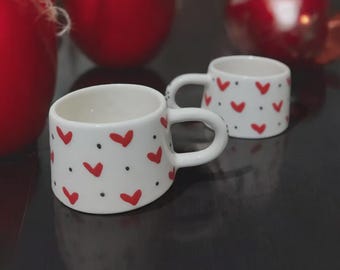 May include: Two white ceramic mugs with red hearts and black dots on a black surface.