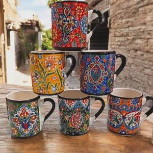 Peut inclure: Six tasses en céramique colorées avec des motifs floraux complexes. Les tasses sont empilées en forme de pyramide sur une table en bois. Les tasses sont toutes de couleurs différentes, notamment rouge, bleu, jaune, vert et orange.