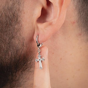 Può includere: Orecchino color argento con un pendente a forma di croce. La croce presenta dettagli decorativi ed è sospesa da un orecchino a cerchio. L'orecchino è indossato all'orecchio.