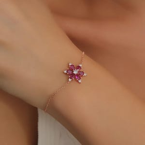 May include: Rose gold bracelet with a flower-shaped pendant. The pendant features ruby-coloured gemstones and small white gemstones. The bracelet is delicate and elegant, suitable for special occasions.