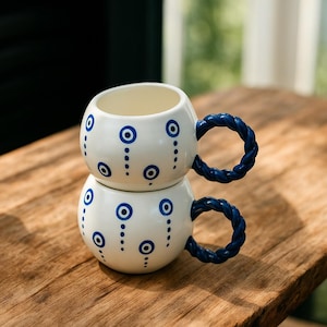 May include: Two white ceramic mugs stacked. Each mug features a blue, braided handle and is decorated with blue eye-shaped designs and dotted lines. The mugs are on a wooden surface.