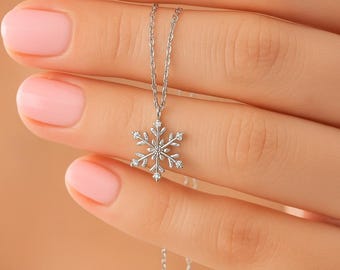 May include: A delicate silver necklace featuring a snowflake pendant with small, sparkling accents. The pendant is suspended from a fine chain. The necklace is held up by a person's hand with pink nail polish.