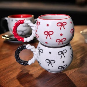 Puede incluir: Dos tazas de cerámica blanca apiladas, cada una con forma redondeada y asa trenzada. La taza superior tiene lazos rojos y borde rojo, mientras que la inferior tiene lazos negros y asa negra. Para café o té.