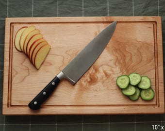 May include: A rectangular wooden cutting board with a groove around the edge. A large chef's knife with a black handle rests on the board, next to sliced apples and cucumbers. The board measures 10 inches by 15 inches.