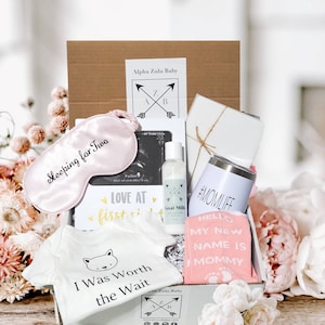 May include: A pink and white baby shower gift box with a variety of items, including a sleep mask, lotion, a tumbler, socks, and a onesie. The box is decorated with the text "Alpha Zulu Baby" and the onesie says "I Was Worth the Wait".