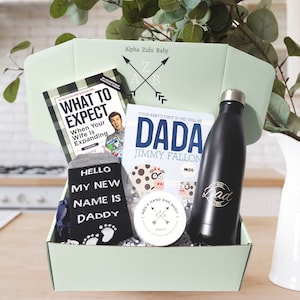 May include: A green gift box with a variety of items for a new dad. The items include a book titled "What to Expect When Your Wife Is Expanding", a book titled "Dada" by Jimmy Fallon, a pair of socks that say "Hello My New Name Is Daddy", a black water bottle with the word "Dad" on it, and a small jar of lotion.
