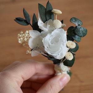 May include: A miniature white floral arrangement with a central rose, delicate white flowers, and green leaves. The arrangement includes small, round, beige accents and is bound at the base with white ribbon.