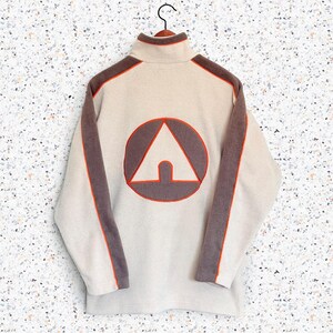 May include: A cream-colored fleece pullover featuring a high collar and long sleeves with dark brown panels running down the arms. The back displays a large circular graphic with a dark brown triangle and a bright orange outline trim.