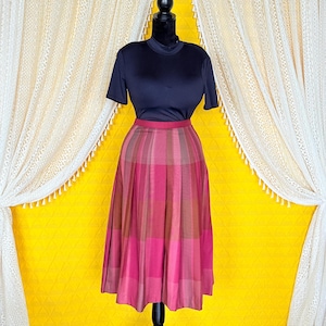 May include: A navy blue short-sleeved top and a pleated midi skirt. The skirt has a plaid pattern in pink, red, and brown. The outfit is displayed on a black mannequin against a yellow backdrop with white lace curtains.