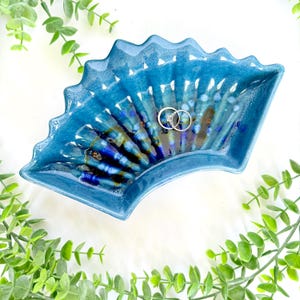 May include: A blue ceramic fan-shaped dish with a textured surface. The dish has a scalloped edge and holds two silver rings. The interior features a blend of blue and brown hues. Greenery surrounds the dish.