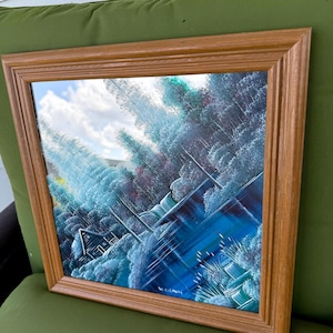 May include: A framed landscape painting featuring a cabin nestled among blue and white trees, with a blue river and cloudy sky. The painting is in a wooden frame.