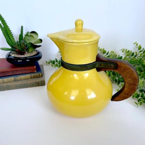 May include: A yellow ceramic coffee pot with a wooden handle and a lid. The pot has a rounded base and a spout for pouring. A dark green band encircles the pot. The handle is made of dark wood.