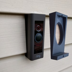 May include: A black Ring doorbell with a white "ring" logo is mounted on a white siding. A black plastic mounting bracket is also shown.