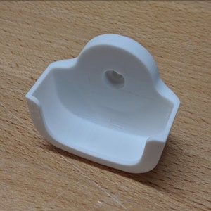 May include: White plastic wall mount with a rounded top and a small hole in the center. The mount has a curved, open space on the bottom.