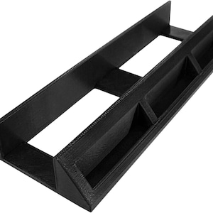 May include: A black, rectangular desk organizer with multiple compartments. The organizer has a long, open central section flanked by smaller, angled storage spaces. The design features a textured surface and is made of a solid, opaque material.