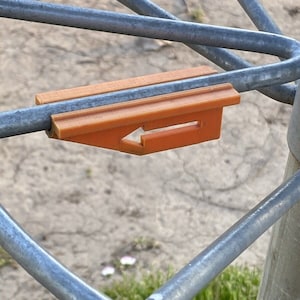 May include: Orange plastic clip with an arrow pointing to the left, attached to a metal fence.