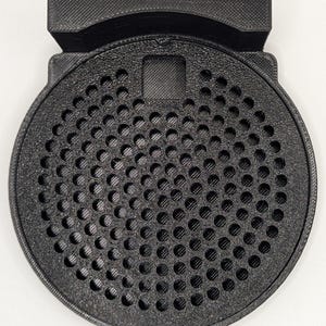 May include: Black plastic speaker grille with a circular design and a small rectangular opening in the center. The grille has a textured surface and is designed to protect the speaker from dust and debris.