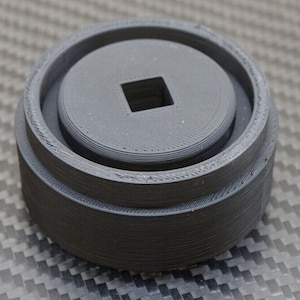 May include: A black plastic 3D printed tool with a square hole in the center. The tool has a raised lip around the edge.