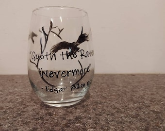 May include: A clear stemless wine glass with black artwork and text. The design includes a raven, tree branches, and the quote "Quoth the Raven 'Nevermore'" by Edgar Allan Poe. The glass is on a gray surface.